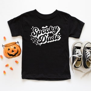 May include: Black t-shirt with white text that says "Spooky Dude" with a spider web and bat graphic. The shirt is laid flat on a white surface with a small orange pumpkin and candy corn scattered around it. There are also black sneakers with white laces on the right side of the shirt.