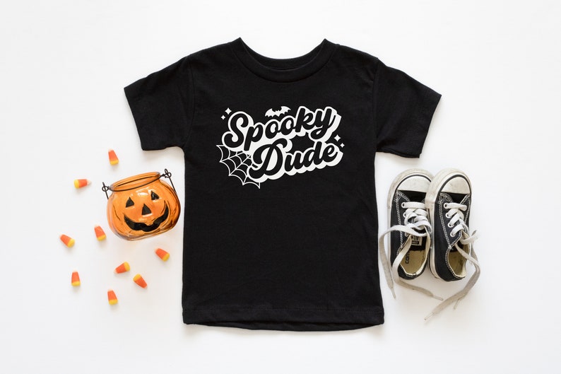May include: Black t-shirt with white text that says "Spooky Dude" with a spider web and bat graphic. The shirt is laid flat on a white surface with a small orange pumpkin and candy corn scattered around it. There are also black sneakers with white laces on the right side of the shirt.