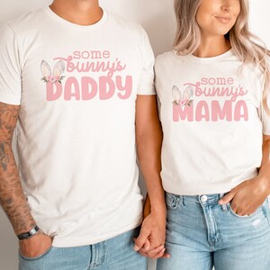 May include: Two white t-shirts with pink text and bunny ears. The text on the left shirt says "Some Bunny's Daddy" and the text on the right shirt says "Some Bunny's Mama".
