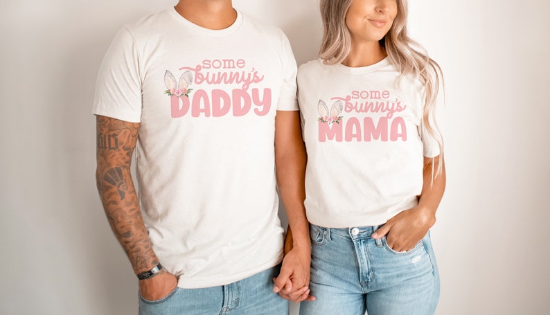 May include: Two white t-shirts with pink text and bunny ears. The text on the left shirt says "Some Bunny's Daddy" and the text on the right shirt says "Some Bunny's Mama".