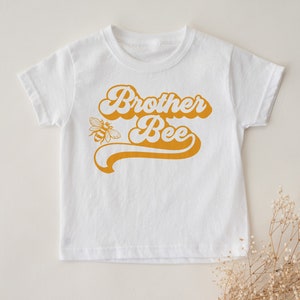 Pu&ograve; includere: Maglietta bianca a maniche corte con una grafica gialla in stile retr&ograve; che dice "Brother Bee" e una piccola illustrazione di un'ape.