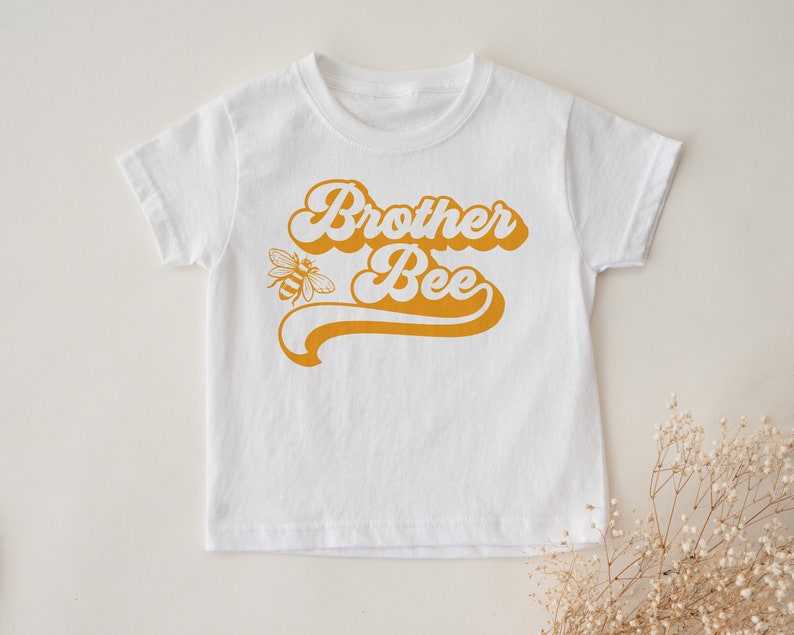 Pu&ograve; includere: Maglietta bianca a maniche corte con una grafica gialla in stile retr&ograve; che dice "Brother Bee" e una piccola illustrazione di un'ape.