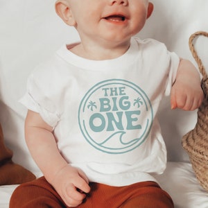 May include: A white t-shirt with a blue circle graphic that says "The Big One" with palm trees and a wave. The shirt is on a baby wearing brown pants.