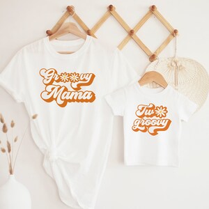 May include: Two white t-shirts with a retro font design. The larger shirt says "Groovy Mama" with a daisy in the word "Groovy". The smaller shirt says "Two Groovy" with a daisy in the word "Two".