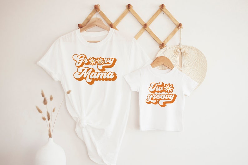 May include: Two white t-shirts with a retro font design. The larger shirt says "Groovy Mama" with a daisy in the word "Groovy". The smaller shirt says "Two Groovy" with a daisy in the word "Two".