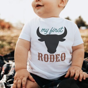 May include: A white baby onesie with a black bull silhouette and the text "my first rodeo" in brown and teal lettering.