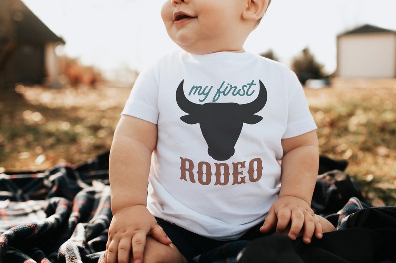 May include: A white baby onesie with a black bull silhouette and the text "my first rodeo" in brown and teal lettering.
