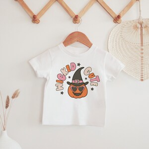 Puede incluir: Camiseta blanca con un gr&aacute;fico de una calabaza con un sombrero de bruja y gafas de sol. El texto "Wicked Cute" est&aacute; escrito alrededor de la calabaza.