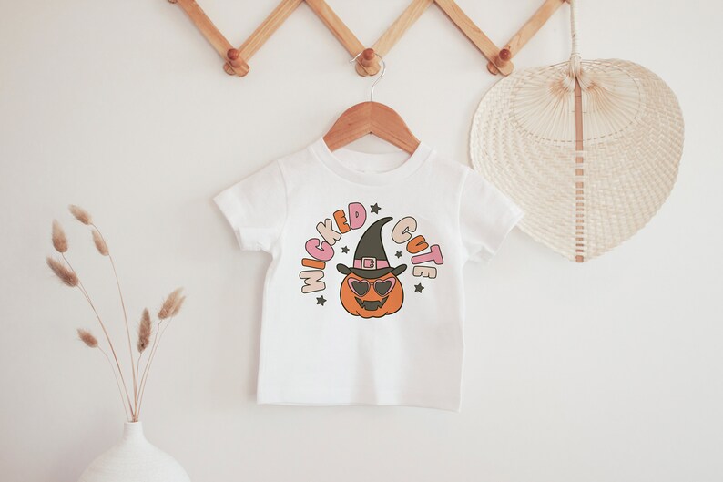 Puede incluir: Camiseta blanca con un gr&aacute;fico de una calabaza con un sombrero de bruja y gafas de sol. El texto "Wicked Cute" est&aacute; escrito alrededor de la calabaza.