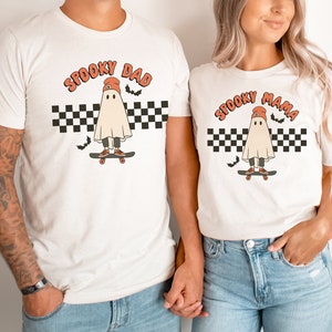 May include: Two white t-shirts with a ghost wearing a red beanie and skateboarding on a checkered background. The text on the shirts reads "Spooky Dad" and "Spooky Mama".