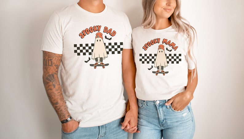 May include: Two white t-shirts with a ghost wearing a red beanie and skateboarding on a checkered background. The text on the shirts reads "Spooky Dad" and "Spooky Mama".