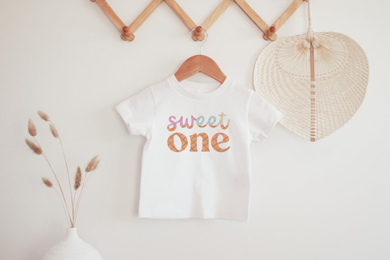 May include: White baby onesie with the text "sweet one" in colorful letters with a patterned background.
