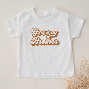 May include: White t-shirt with brown text that says "Groovy Brother" with a daisy graphic.