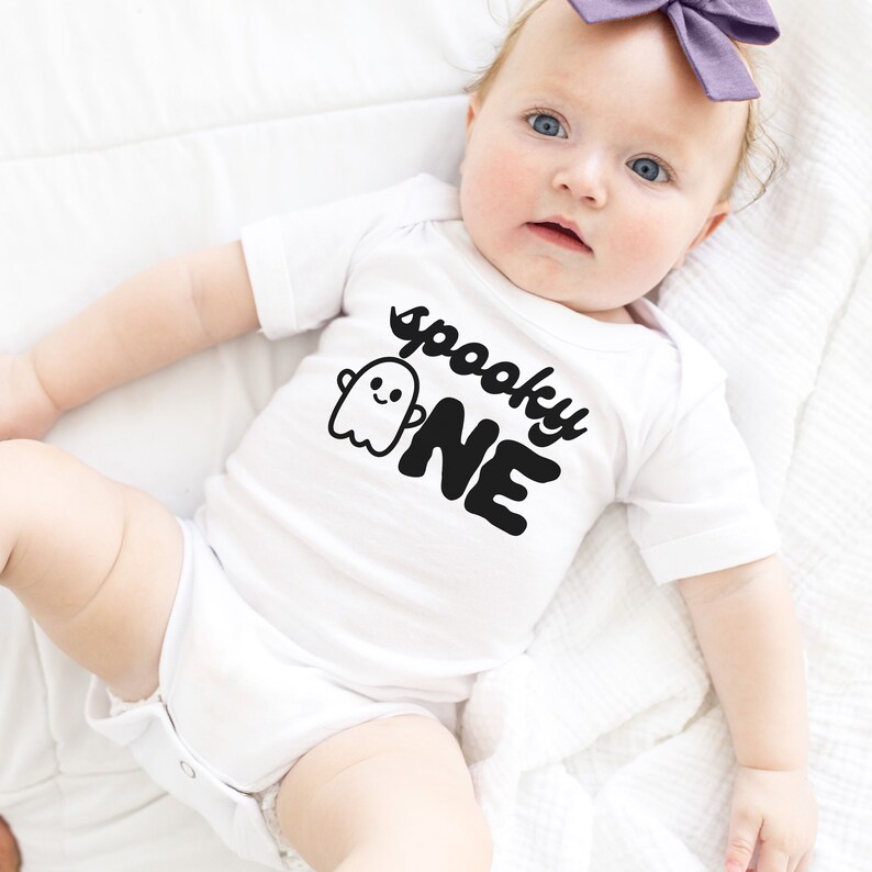 May include: A white baby onesie with a black ghost and the text "spooky one" printed on the front.