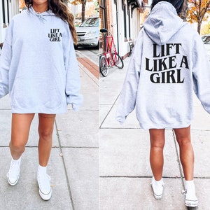 K&ouml;nnte beinhalten: Grauer Kapuzenpullover mit dem Aufdruck "Lift Like A Girl" in Schwarz auf der R&uuml;ckseite. Das Model tr&auml;gt den Pullover mit wei&szlig;en Turnschuhen.
