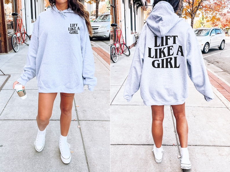 K&ouml;nnte beinhalten: Grauer Kapuzenpullover mit dem Aufdruck "Lift Like A Girl" in Schwarz auf der R&uuml;ckseite. Das Model tr&auml;gt den Pullover mit wei&szlig;en Turnschuhen.