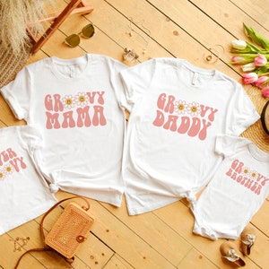 K&ouml;nnte beinhalten: Eine Auswahl an wei&szlig;en T-Shirts und ein Baby-Body mit rosa Retro-Text und G&auml;nsebl&uuml;mchen-Grafiken. Die Shirts zeigen "Groovy Mama", "Groovy Daddy", "4ever Groovy" und "Groovy Brother". Ideal f&uuml;r eine Familie, auf einer Holzoberfl&auml;che angeordnet.