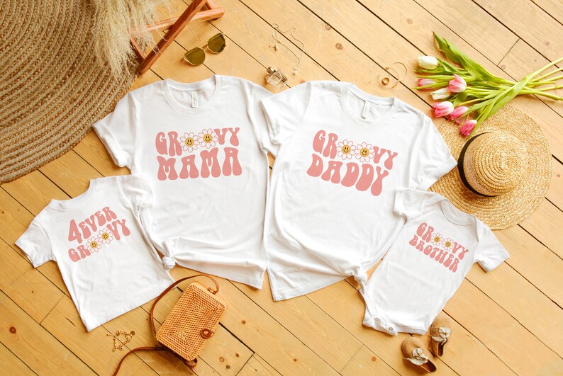 K&ouml;nnte beinhalten: Eine Auswahl an wei&szlig;en T-Shirts und ein Baby-Body mit rosa Retro-Text und G&auml;nsebl&uuml;mchen-Grafiken. Die Shirts zeigen "Groovy Mama", "Groovy Daddy", "4ever Groovy" und "Groovy Brother". Ideal f&uuml;r eine Familie, auf einer Holzoberfl&auml;che angeordnet.
