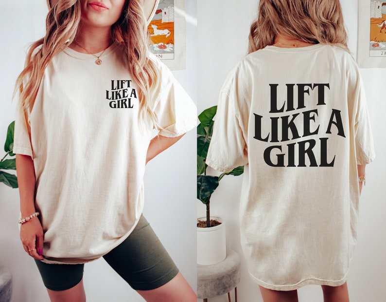 K&ouml;nnte beinhalten: Ein cremefarbenes T-Shirt mit dem Text "Lift Like A Girl" in Schwarz auf Vorder- und R&uuml;ckseite.