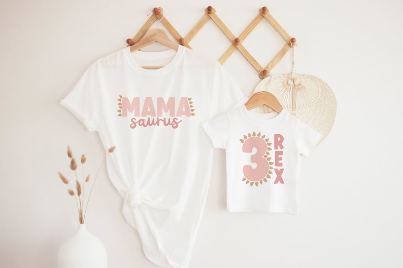 May include: Two white t-shirts with pink and gold dinosaur-themed designs. The larger shirt says "Mama saurus" and the smaller shirt says "Rex" with the number "3" in a sun shape.