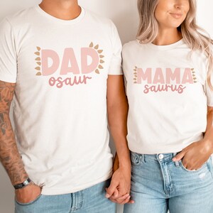 May include: A couple wearing matching white t-shirts with pink and brown text. The man's shirt says "DAD-osaur" and the woman's shirt says "MAMA-saurus".