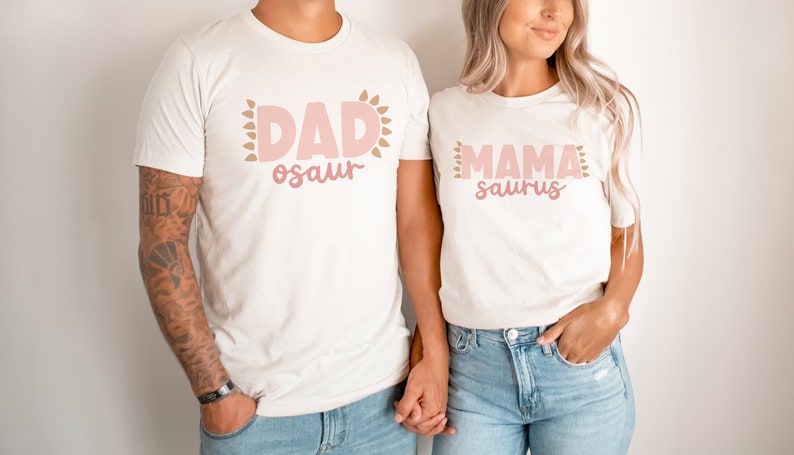 May include: A couple wearing matching white t-shirts with pink and brown text. The man's shirt says "DAD-osaur" and the woman's shirt says "MAMA-saurus".