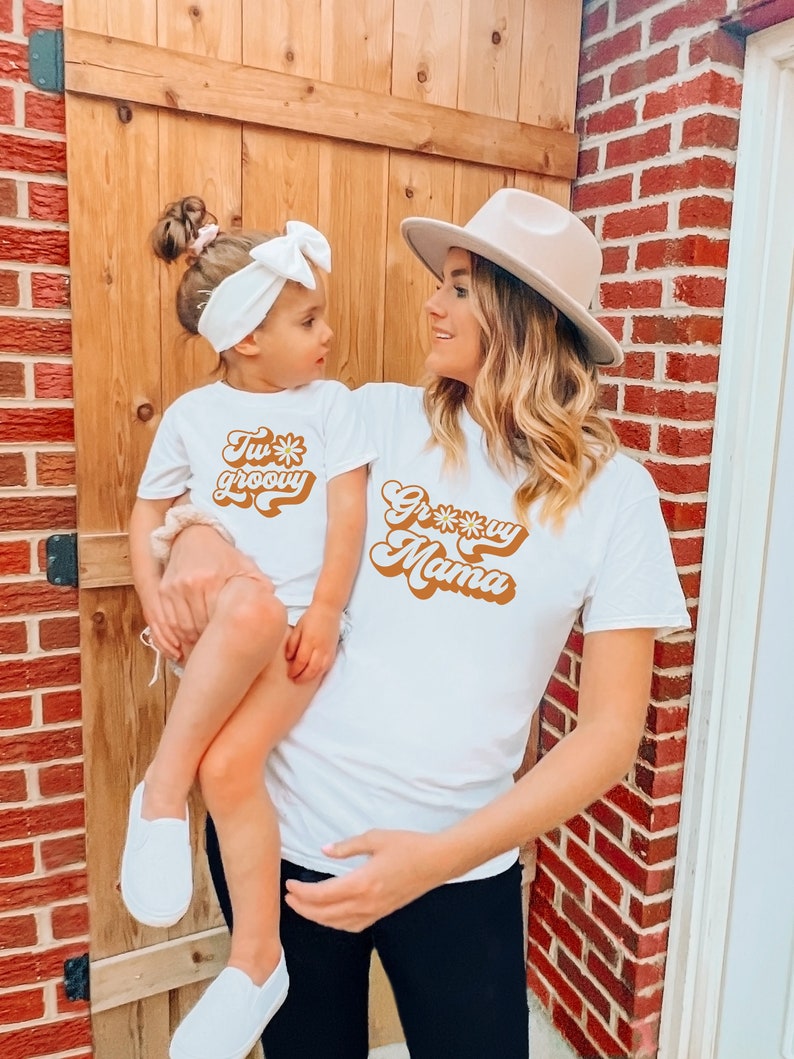 May include: A mother and daughter wearing matching white t-shirts with a retro groovy font. The daughter's shirt says "Too groovy" and the mother's shirt says "Groovy Mama".
