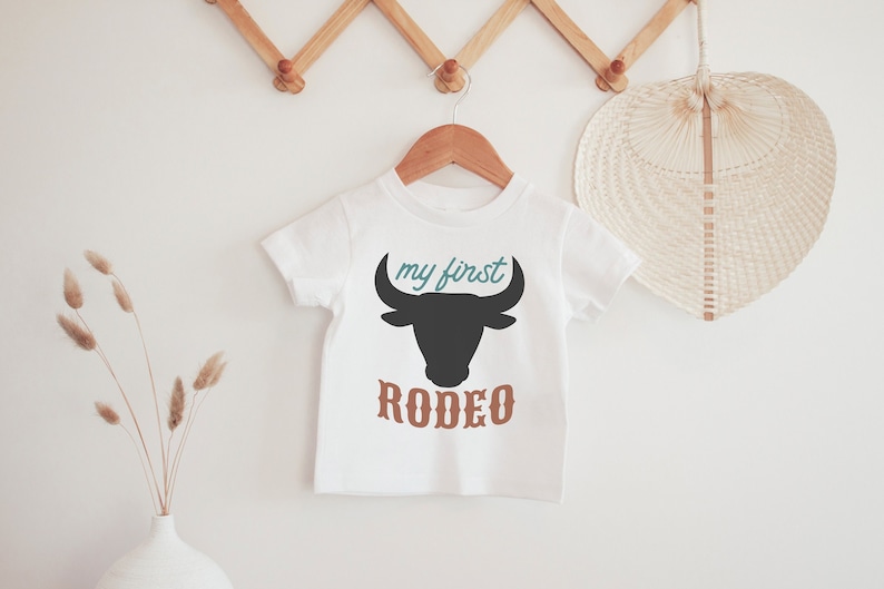May include: White baby t-shirt with a black bull silhouette and the text "my first rodeo" in brown and teal.