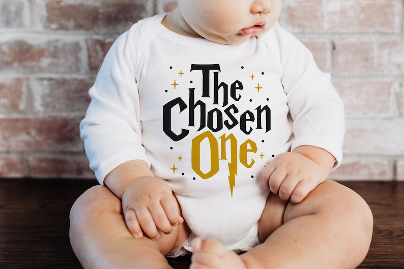 May include: A white long-sleeved onesie with the text "The Chosen One" in black and gold lettering. A lightning bolt is in the gold lettering.