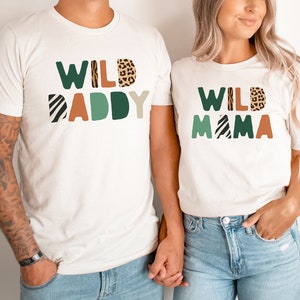May include: Two white t-shirts with the text "WILD DADDY" and "WILD MAMA" in colorful block letters with animal print patterns. The shirts are worn by two people holding hands.