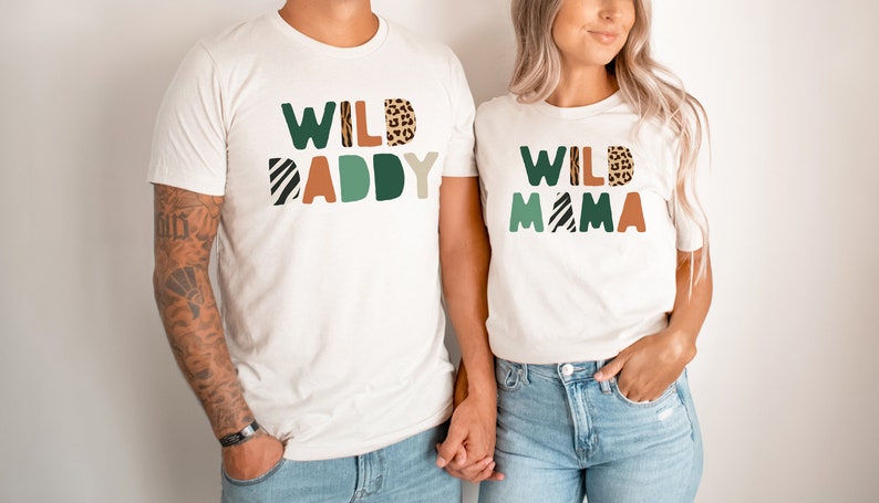 May include: Two white t-shirts with the text "WILD DADDY" and "WILD MAMA" in colorful block letters with animal print patterns. The shirts are worn by two people holding hands.