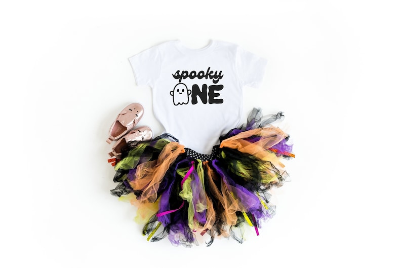 May include: White onesie with a black ghost and the text "spooky one" on it. A multi-colored tulle tutu and pink shoes are also pictured.