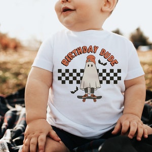 May include: White baby onesie with a graphic of a ghost wearing a red beanie and skateboarding. The text "Birthday Dude" is written in orange above the ghost. The background is a black and white checkered pattern with bats.