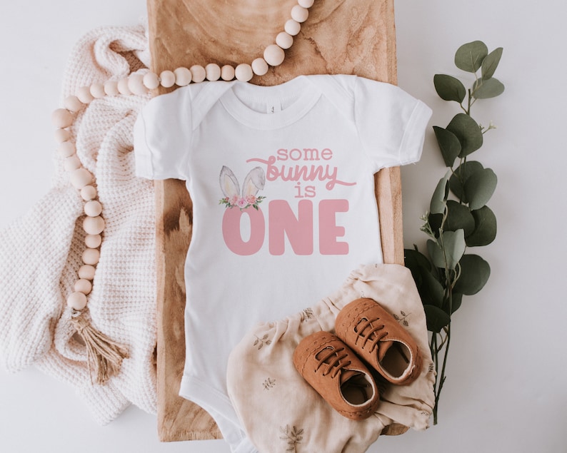 May include: White baby onesie with pink bunny ears and the text "some bunny is one" in pink lettering.