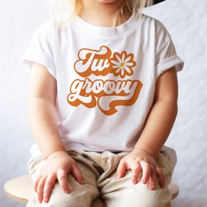 May include: A white t-shirt with a retro-style orange graphic that says "Two groovy" with a white daisy.