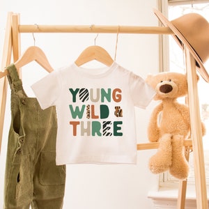 May include: White t-shirt with the text "YOUNG WILD & THREE" in colorful, patterned letters. The shirt is hanging on a wooden rack with a pair of green overalls and a stuffed teddy bear.