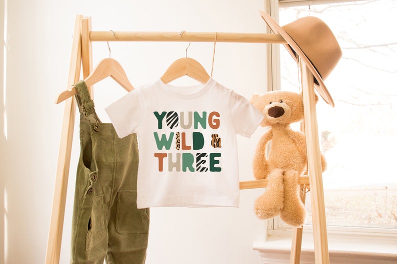 May include: White t-shirt with the text "YOUNG WILD & THREE" in colorful, patterned letters. The shirt is hanging on a wooden rack with a pair of green overalls and a stuffed teddy bear.