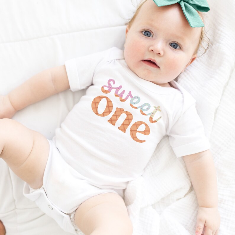 May include: A white onesie with the words "sweet one" in colorful letters with a waffle cone pattern. The onesie is worn by a baby with a green bow in their hair.