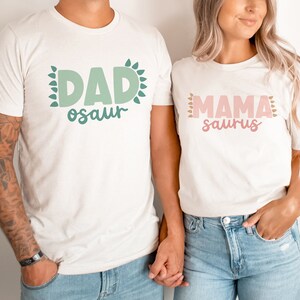 May include: A couple wearing white t-shirts with the text "DAD-osaur" and "MAMA-saurus" in green and pink lettering. The shirts have a leafy design around the text.