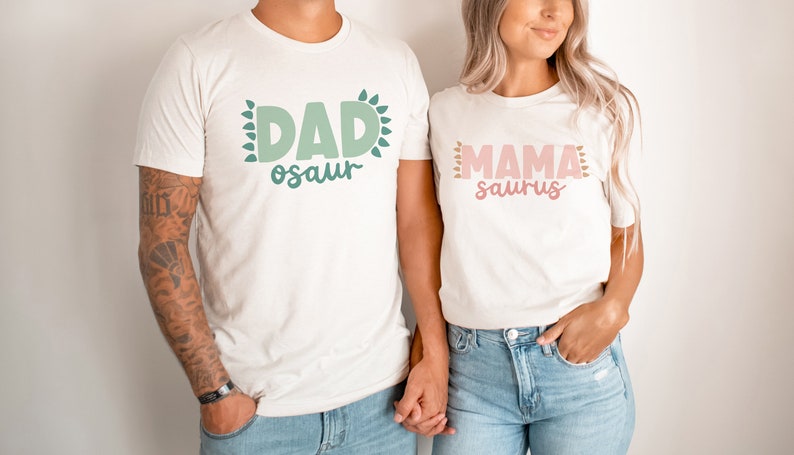 May include: A couple wearing white t-shirts with the text "DAD-osaur" and "MAMA-saurus" in green and pink lettering. The shirts have a leafy design around the text.