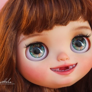 May include: Close-up of a doll's face with green eyes, brown hair, and a toothy smile. The doll has a pink blush on its cheeks and a light pink mouth.