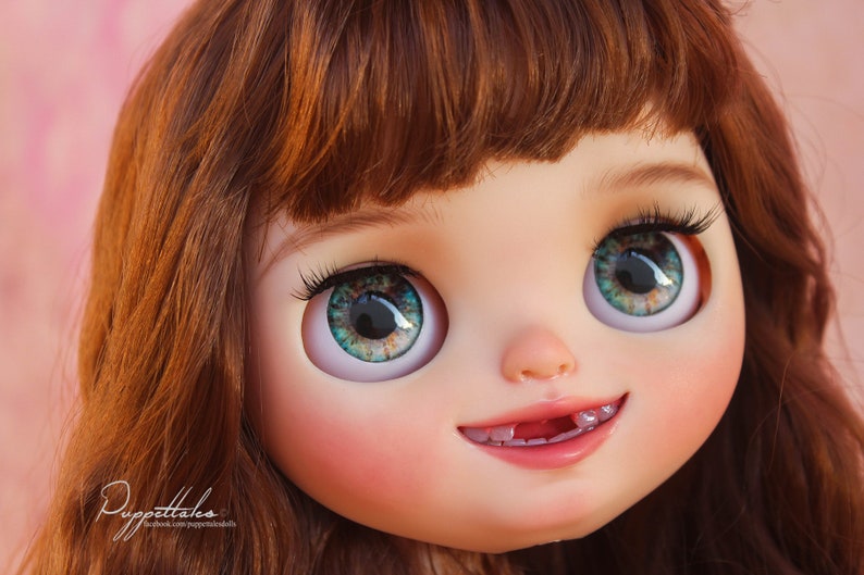 May include: Close-up of a doll's face with green eyes, brown hair, and a toothy smile. The doll has a pink blush on its cheeks and a light pink mouth.
