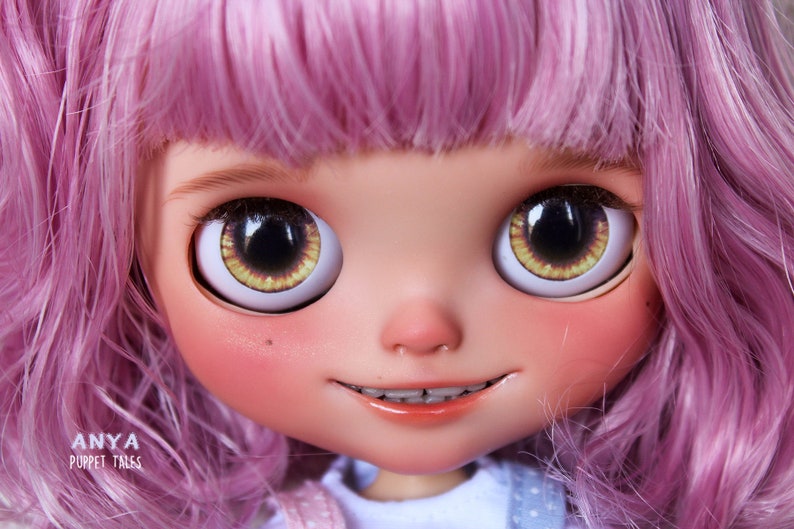 May include: A close-up of a doll's face with pink hair, large brown eyes with yellow irises, and a slight smile. The doll is wearing braces and has rosy cheeks. The text 'ANYA PUPPET TALES' is visible at the bottom of the image.