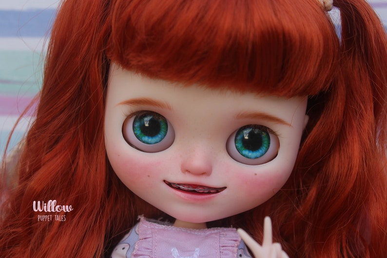 May include: A doll with red hair, blue eyes, and a pink and gray dress. The doll is smiling and has braces on her teeth. The text 'Willow Puppet Tales' is visible at the bottom of the image.