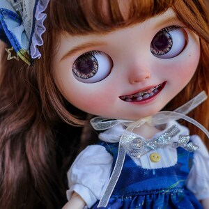 May include: A doll with long brown hair wearing a blue and white dress with a silver ribbon. The doll has blue eyes and is smiling. The doll is wearing braces.