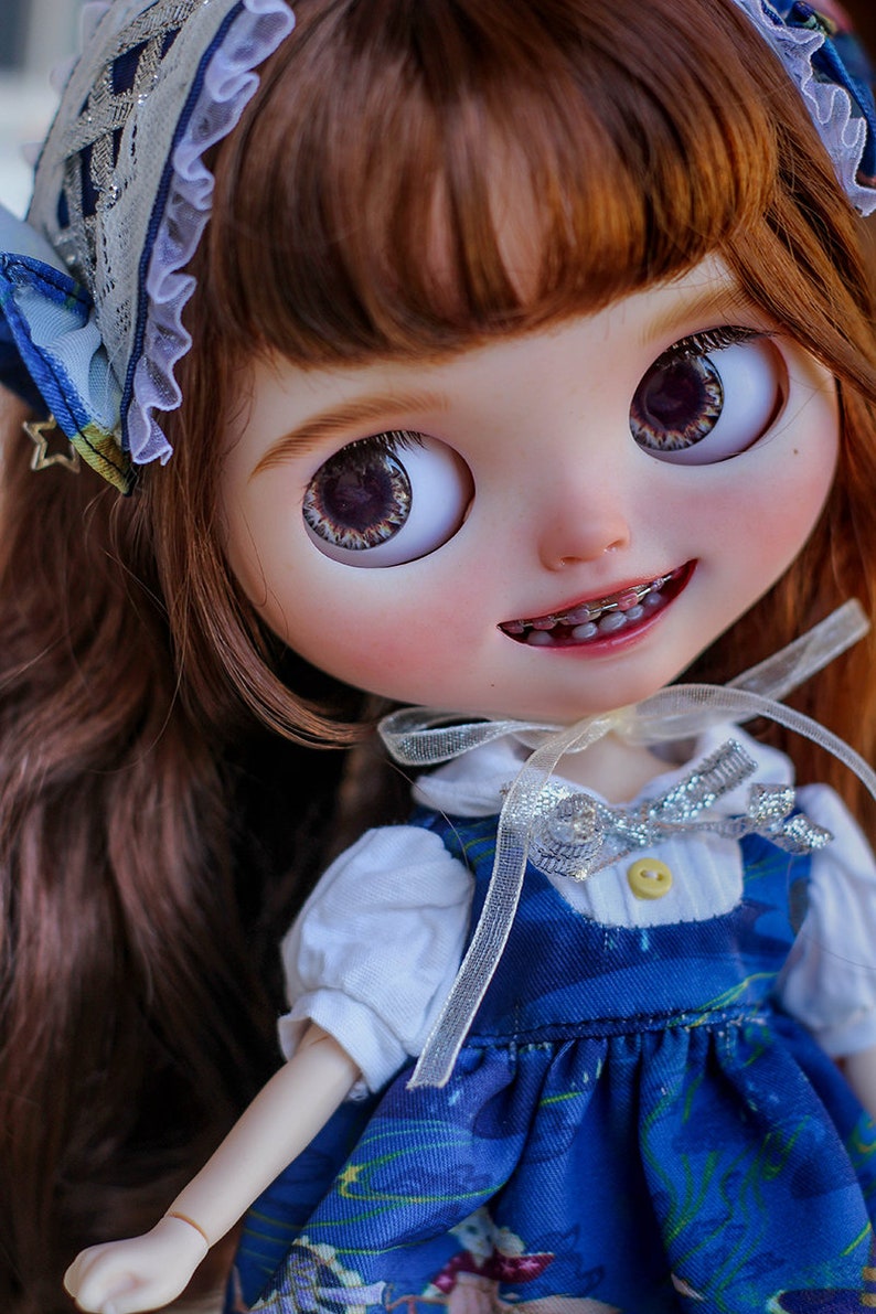 May include: A doll with long brown hair wearing a blue and white dress with a silver ribbon. The doll has blue eyes and is smiling. The doll is wearing braces.