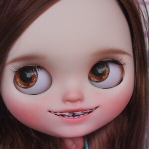 May include: A doll with brown hair and brown eyes. The doll is smiling and has braces on its teeth.