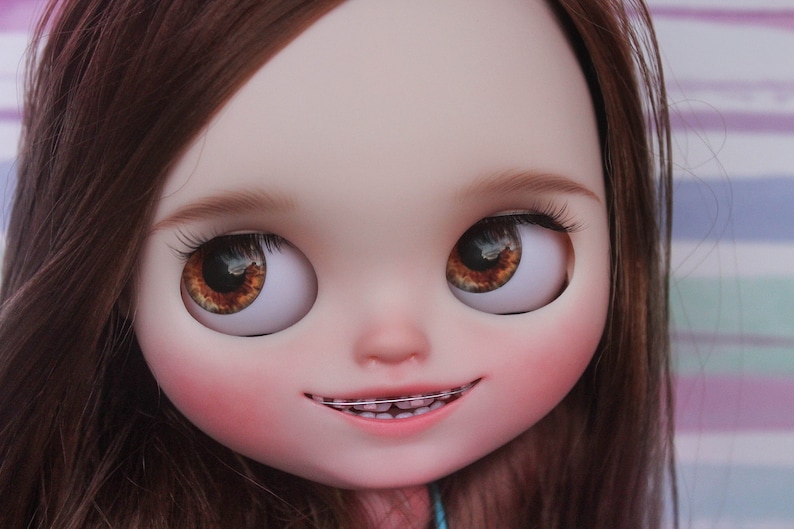 May include: A doll with brown hair and brown eyes. The doll is smiling and has braces on its teeth.
