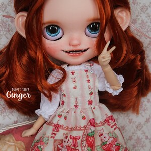 May include: A red-haired doll wearing a pink and white dress with a strawberry and bunny print. The dress has a white ruffle trim. The doll has blue eyes and is smiling. The text 'Puppet Tales Ginger' is visible on the doll's dress.