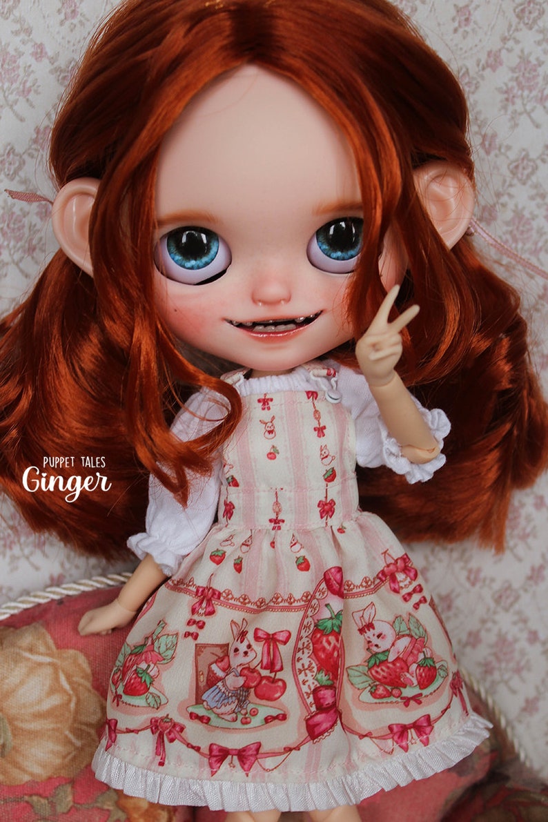 May include: A red-haired doll wearing a pink and white dress with a strawberry and bunny print. The dress has a white ruffle trim. The doll has blue eyes and is smiling. The text 'Puppet Tales Ginger' is visible on the doll's dress.